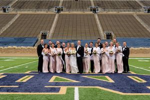 Bride and groom and their wedding party posing for a picture on the field. The bridesmaids are in light pink dresses and the groomsmen are in black suits.